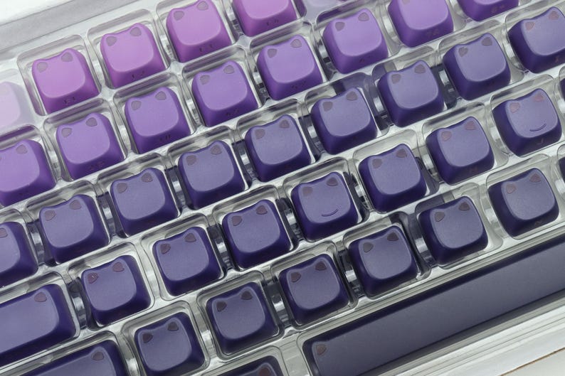 May include: A close-up of a keyboard with custom keycaps. The keycaps are a gradient of purple hues, each featuring a stylized frog face. The keyboard keys are arranged in a grid pattern, with a dark purple frame.