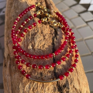 May include: Three layered bracelets with red gemstone beads and gold accents. The bracelets are stacked on a wooden surface.