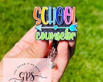 May include: A black glitter badge holder with a rainbow colored "School Counselor" text and a silver arrow. The badge holder is attached to a retractable badge reel.