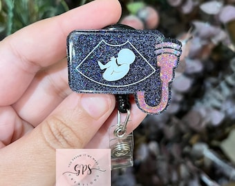 May include: Black glitter badge reel with a white ultrasound image of a baby and a pink and white cord. The badge reel has a silver retractable cord and a small white tag with the text "You Dream It We Make It GPS Creations".
