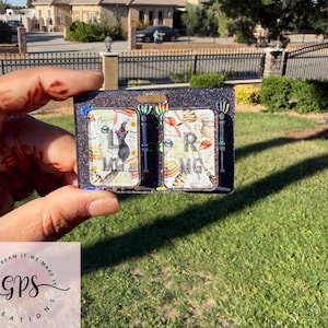 May include: A rectangular black glitter resin badge reel topper featuring two white inserts with a witch hat, a cat, and the letters L, R, M, and G. The design includes skeleton hand accents on the sides and a small rectangular cutout at the top.