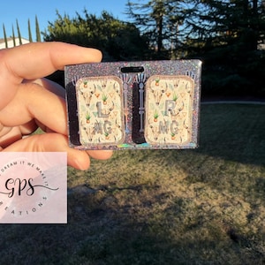 May include: A rectangular badge holder with a glittery, dark gray border. The holder contains two rectangular inserts with a cream background, featuring a steer skull and plant pattern. The inserts are labeled "L MG" and "R MG".