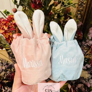May include: Two personalized bunny-shaped drawstring bags, one pink with the name "Marissa" and one blue with the name "Mason". The bags are made of soft velvet and have white bunny ears.