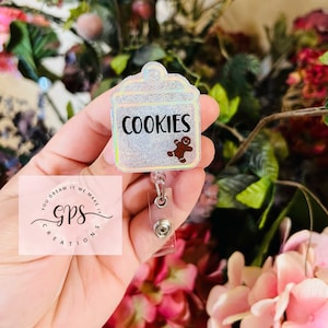 May include: A sparkly silver glitter badge holder with a clear plastic retractable reel. The badge holder has a gingerbread man and the text "COOKIES" printed on it. The badge holder is attached to a clear plastic retractable reel. The badge holder is held in a hand.