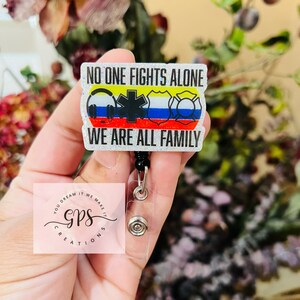 May include: A glittery badge reel with the text "NO ONE FIGHTS ALONE WE ARE ALL FAMILY." The badge features symbols for emergency services and a logo for GPS Creations. The reel is black and has a clear plastic holder.