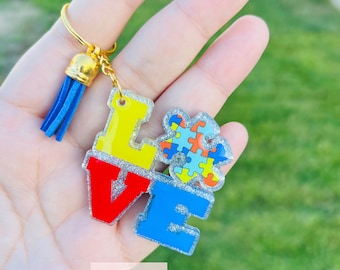 May include: A keychain with the word "LOVE" in red, yellow, and blue letters, with a puzzle piece design. The keychain has a gold-colored ring and a blue tassel. The letters and puzzle piece have a glittery border.