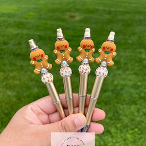 May include: Five gold pens with gingerbread man toppers. Each topper has a red and green bow tie, a white pearl, and a pink and white hat. The pens are held in a hand. The text "You dream it, we make it, GPS Creations" is on a pink and white sticker.