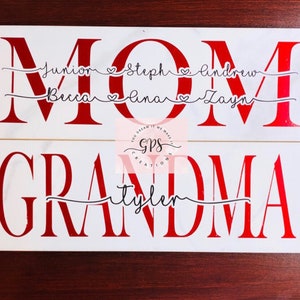 May include: White wooden sign with the word "MOM" in large red letters. Below the word "MOM" are the names "Junior, Steph, Andrew, Breca, Alana, Zayn" in black cursive writing. Below the word "MOM" is a second white wooden sign with the word "GRANDMA" in large red letters. Below the word "GRANDMA" is the name "Tyler" in black cursive writing.