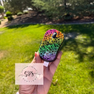 May include: A wireless computer mouse featuring a vibrant rainbow gradient background with a black leopard print pattern. The device includes a black scroll wheel and a small white battery pull tab extending from the bottom edge of the mouse casing.