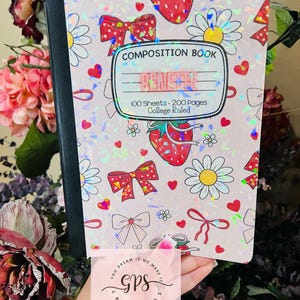 Peut inclure: Un cahier de composition avec une couverture rose orn&eacute;e de n&oelig;uds, de fraises et de marguerites. La couverture porte l'inscription "COMPOSITION BOOK", "RENSHAE" et "100 Sheets &bull; 200 Pages &bull; College Ruled". Le cahier est tenu devant un fond floral.