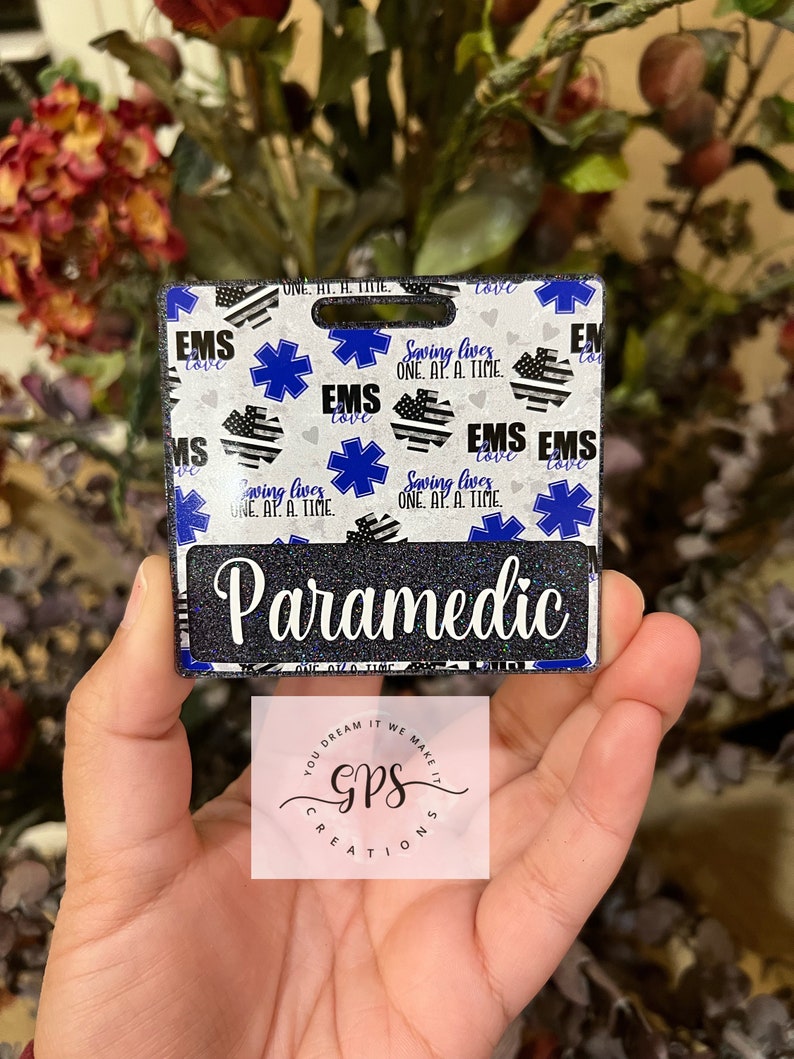 May include: A white and blue patterned badge with the text "EMS" and "Saving Lives One At A Time" repeated. The badge has a black glitter background and the text "Paramedic" in white letters. The badge is held in a hand.