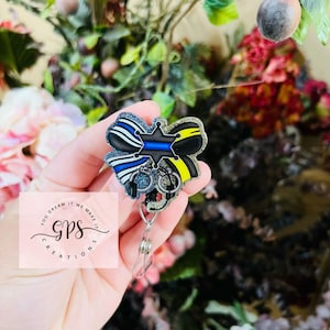 May include: A black and blue thin blue line police badge with a silver star and handcuffs, attached to a clear retractable badge reel. The badge is decorated with glitter and rhinestones.