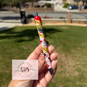 May include: A decorative pen with a red top and a silver tip. The pen's barrel is white with a glittery finish and features various graphics and the text "3 Killer Ross". The pen is held in a hand.