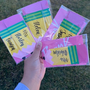 Pu&ograve; includere: Quattro etichette regalo a forma di matita rosa e gialla con strisce verdi. Le etichette sono etichettate con i nomi "Miss Tiamson", "Miss Haslem", "Helen" e "Mrs. Richardson".