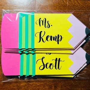 Pu&ograve; includere: Un set di cinque sagome di carta a forma di matita gialla con accenti rosa e verdi. Le sagome hanno il testo "Ms. Kemp is Scott" scritto in nero.