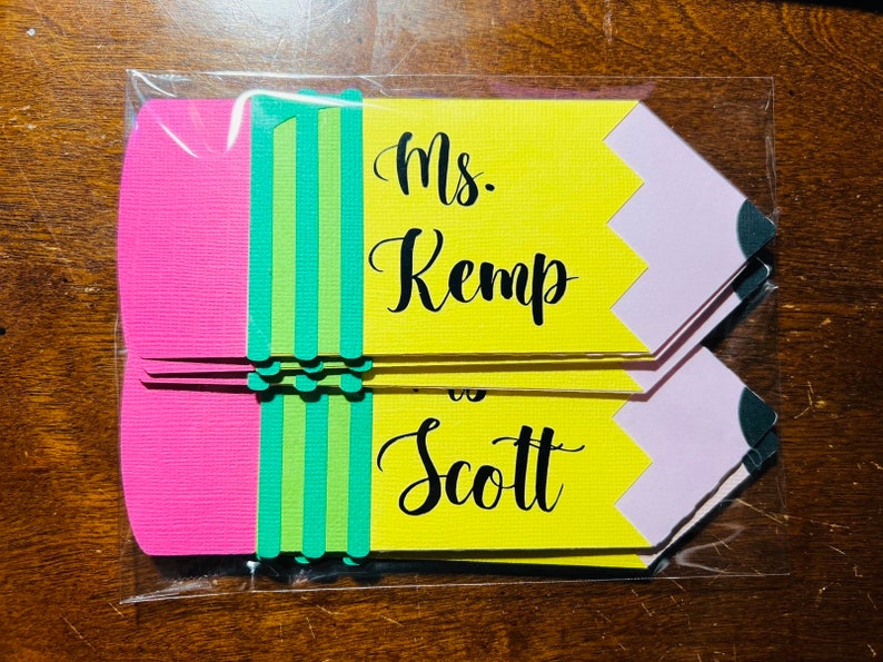Pu&ograve; includere: Un set di cinque sagome di carta a forma di matita gialla con accenti rosa e verdi. Le sagome hanno il testo "Ms. Kemp is Scott" scritto in nero.