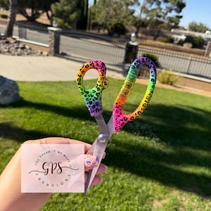 May include: A pair of scissors with a vibrant rainbow-colored leopard print pattern on the handles. The text "LEOPARD AND LACE CO" is printed on the larger handle. The metal blades are silver, and the pivot point features a circular screw.