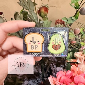 May include: Two enamel pins on a glittery background. One pin is a slice of toast with the letters "L" and "BP". The other pin is an avocado with the letters "R" and "BP". The pins are held in a hand, surrounded by flowers.