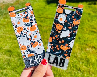 May include: Two Halloween-themed keychains with a glittery finish. Both keychains feature a black and white background with orange pumpkins, ghosts, and bats. The text "LAB" is printed on both keychains.