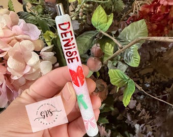 May include: A white glitter pen with the name "DENISE" in red lettering. The pen features red and green bow accents. The pen is held in front of a floral background.