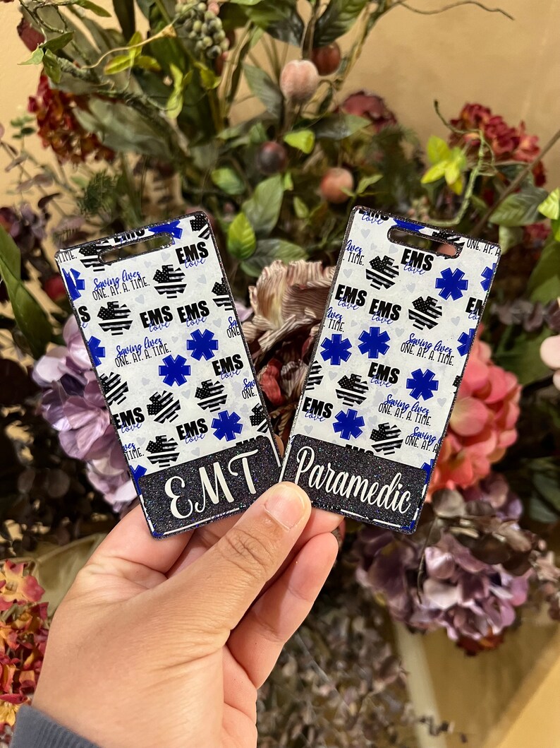May include: Two black and white ID badge holders with glitter. One says "EMT" and the other says "Paramedic". Both have a blue and white star of life and the text "Saving Lives One At A Time".