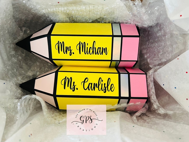 May include: Two yellow pencil-shaped boxes with pink and black accents. The boxes are decorated with the names "Mrs. Micham" and "Ms. Carlisle".