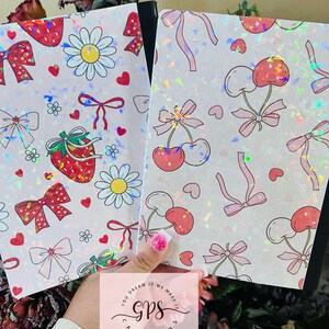 Peut inclure: Deux cahiers avec des couvertures blanches orn&eacute;es de fraises, de marguerites, de cerises et de n&oelig;uds. Les couvertures ont un effet holographique et scintillant. Les cahiers sont tenus devant un fond floral. Le texte "YOU DREAM IT WE MAKE IT GPS CREATIONS" est visible.