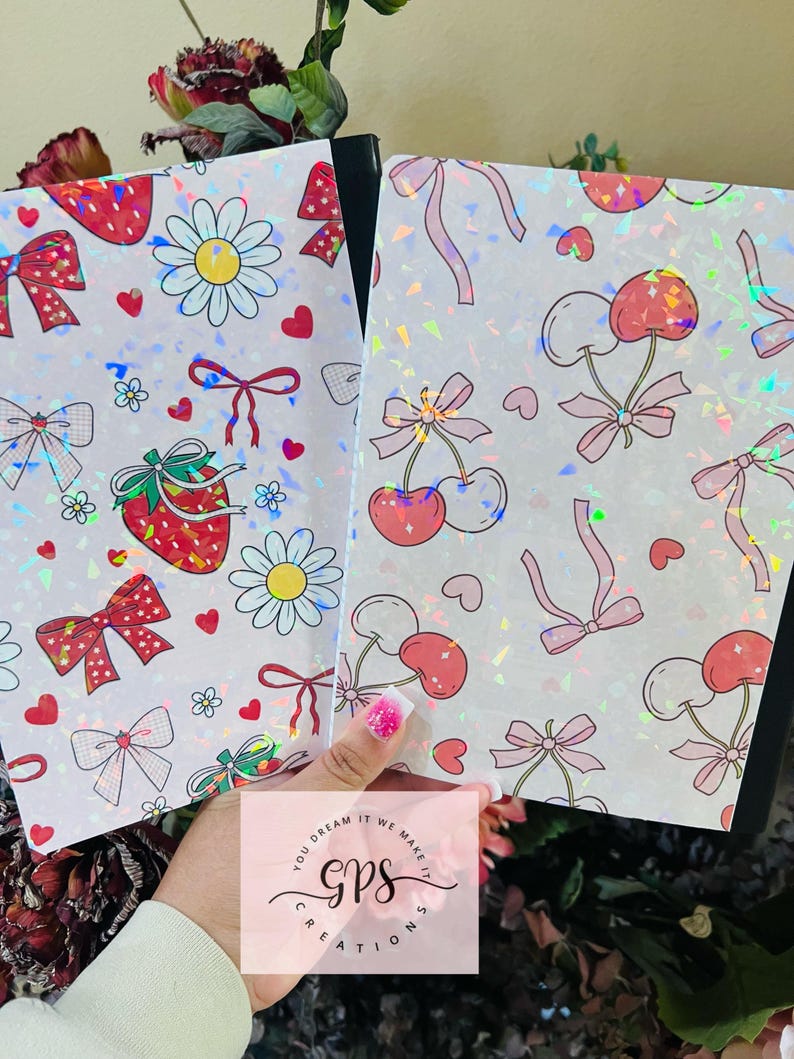 Peut inclure: Deux cahiers avec des couvertures blanches orn&eacute;es de fraises, de marguerites, de cerises et de n&oelig;uds. Les couvertures ont un effet holographique et scintillant. Les cahiers sont tenus devant un fond floral. Le texte "YOU DREAM IT WE MAKE IT GPS CREATIONS" est visible.