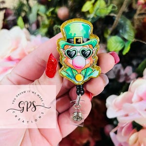May include: A green and gold glitter leprechaun with a pink bubble gum, wearing sunglasses and a green hat, is attached to a black retractable badge reel. The badge reel is in the hand of a person. The text "You Dream It We Make It GPS Creations" is visible in the background.