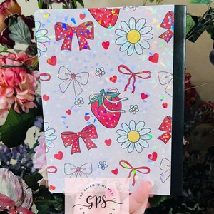 Peut inclure: Un cahier avec une couverture holographique orn&eacute;e de motifs de fraises, de n&oelig;uds, de marguerites et de c&oelig;urs. La couverture a un dos noir et le texte "You dream it we make it GPS Creations". Le cahier est tenu dans une main.