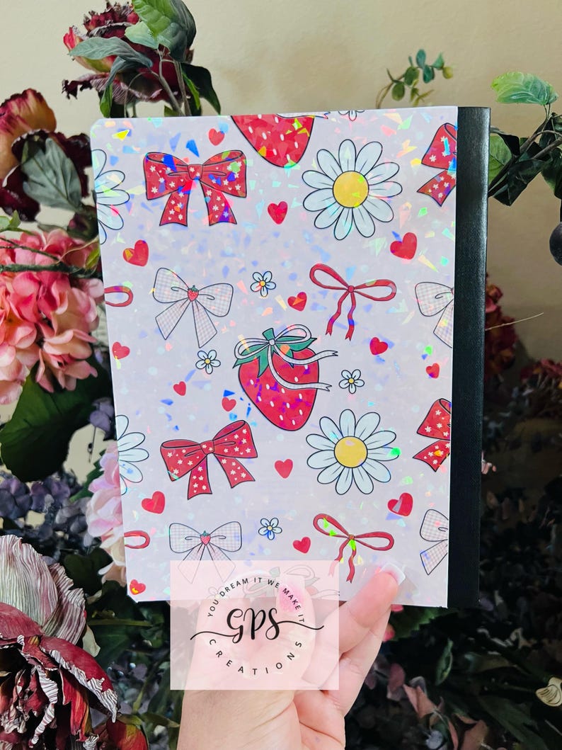 Peut inclure: Un cahier avec une couverture holographique orn&eacute;e de motifs de fraises, de n&oelig;uds, de marguerites et de c&oelig;urs. La couverture a un dos noir et le texte "You dream it we make it GPS Creations". Le cahier est tenu dans une main.