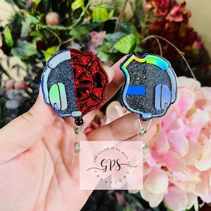 May include: Two black and iridescent badge holders with a red and black firefighter badge and a blue and black police badge. The badges have a headphone design on the side. The badge holders are attached to retractable badge reels. The badge reels are attached to a white background with the text "You dream it, we make it. GPS Creations."