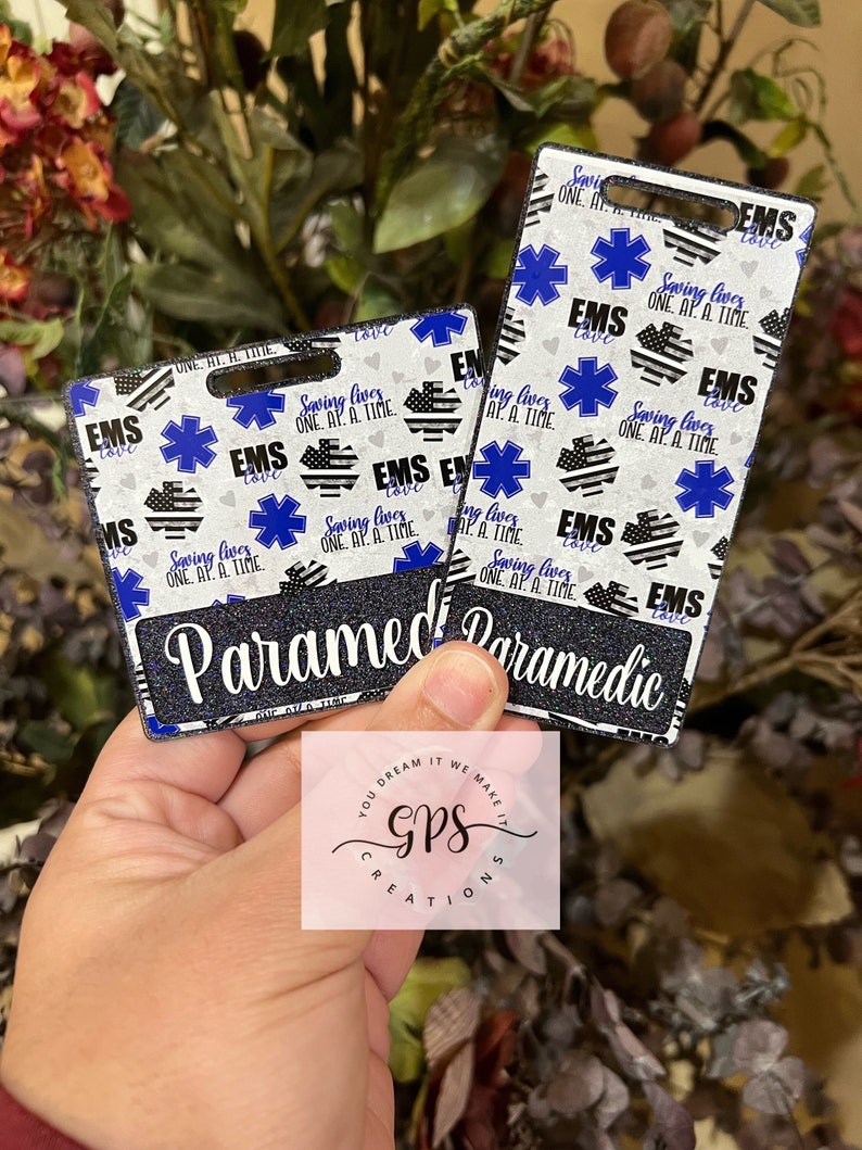 May include: Two black and white patterned ID badge holders with a blue star of life and the text "EMS Love" and "Saving Lives One At A Time". The text "Paramedic" is printed on the bottom of the badge holders.