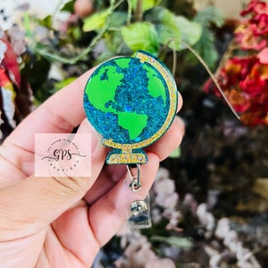 May include: A green and blue glitter globe with a gold rim, attached to a clear retractable badge holder. The globe is on a gold stand. The text "You Dream It We Make It GPS Creations" is visible in the background.