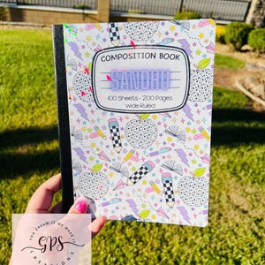 May include: A composition book with a white cover featuring a colorful pattern of apples, pencils, stars, and lightning bolts. The book has the text "COMPOSITION BOOK SANDHU 100 Sheets 200 Pages Wide Ruled". The book is held in a hand.