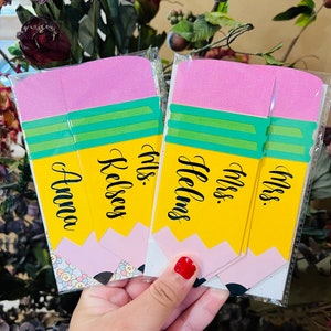 Pu&ograve; includere: Tre carte gialle a forma di matita con gomme rosa. Le carte hanno il testo "Miss. Hellens", "Miss. Kelsey" e "Miss. Anna" scritto in corsivo nero.