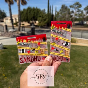 May include: Two red rectangular keychains with a crime scene theme. The keychains feature the text "Killer Found!" and "Do Not Cross" with crime scene elements like a knife, handcuffs, and evidence markers. The name "SANDHU" is printed on the bottom.