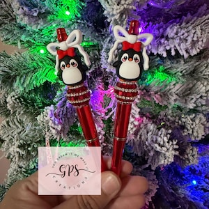 May include: Two red pens with penguin toppers. Each pen features a black and white penguin with a red bow and a decorative red and silver band. The pens are held in front of a Christmas tree with white snow and colorful lights.