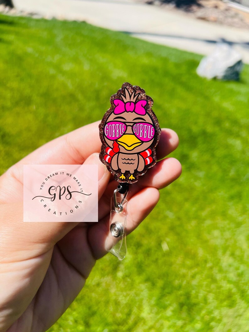 May include: A brown turkey-shaped badge reel with a pink bow and sunglasses that read "GOBBLE GOBBLE." The badge reel has a clear plastic holder and a black retractable cord. The background is green grass.