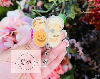 Peut inclure: Un porte-badge avec deux motifs de bébé pour Halloween. Un bébé est déguisé en citrouille, l'autre en momie. Le porte-badge porte les mots "Emergency Department" et un dessin de chauve-souris. Le porte-badge est tenu dans une main.