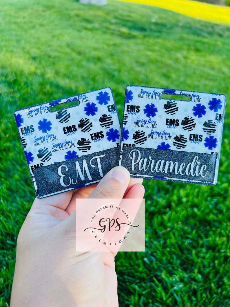 May include: Two black and white patterned keychains with a blue star of life design. The keychains are labeled "EMT" and "Paramedic". The keychains are made by GPS Creations.