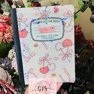 Peut inclure: Un cahier de composition avec une couverture holographique orn&eacute;e de cerises roses, de n&oelig;uds et de c&oelig;urs. La couverture indique "RENEGAE" et "COMPOSITION BOOK" avec des d&eacute;tails sur le nombre de feuilles et le lignage. Le cahier est tenu devant un fond floral.