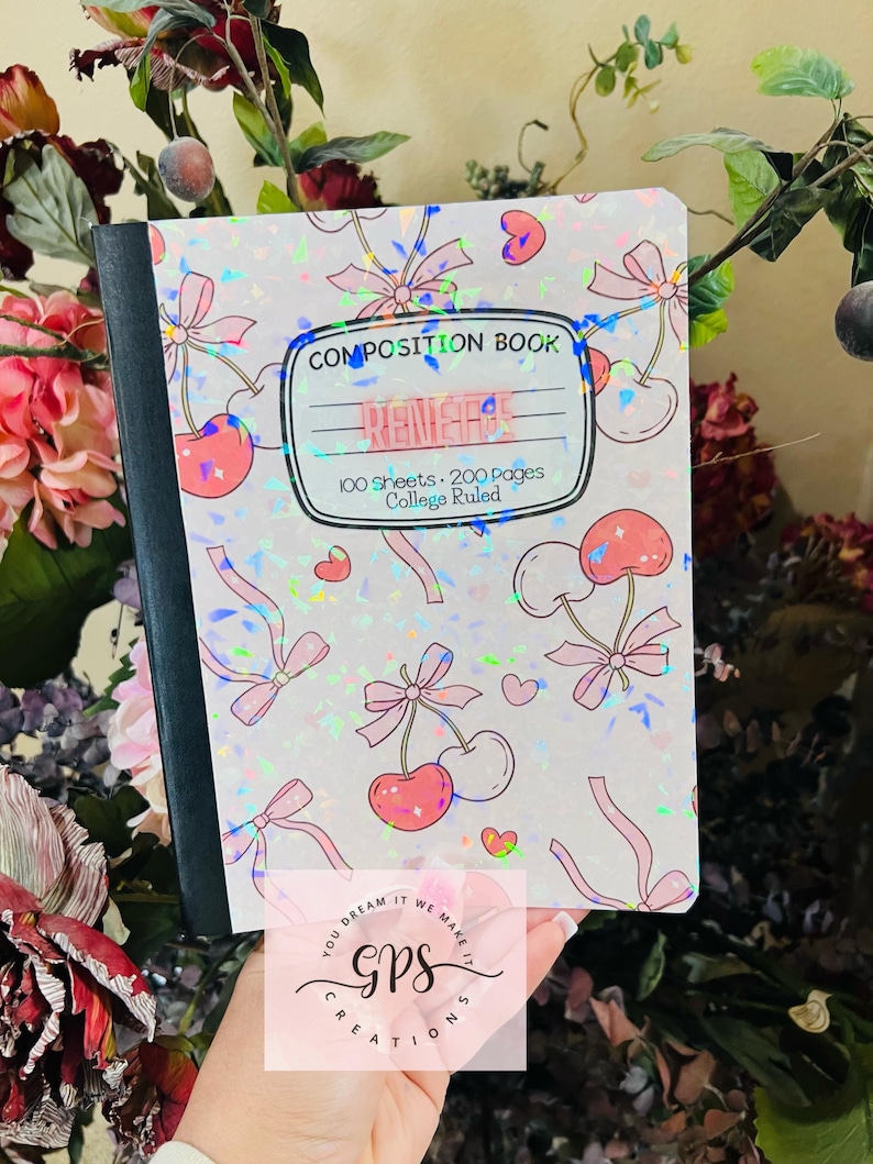 Peut inclure: Un cahier de composition avec une couverture holographique orn&eacute;e de cerises roses, de n&oelig;uds et de c&oelig;urs. La couverture indique "RENEGAE" et "COMPOSITION BOOK" avec des d&eacute;tails sur le nombre de feuilles et le lignage. Le cahier est tenu devant un fond floral.