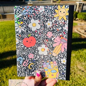 May include: A composition notebook with a black and white speckled pattern, decorated with colorful school-themed stickers. The stickers include a smiling sun, flowers, an apple, a pencil, and the letters A, B, and C.