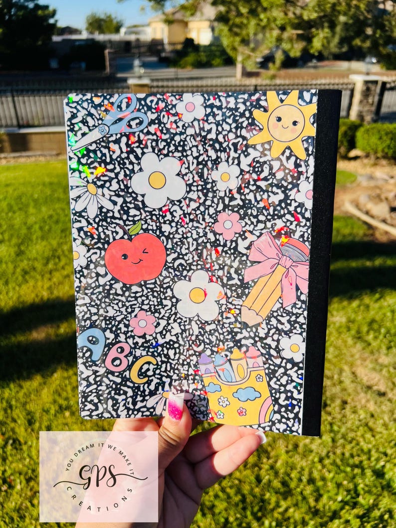 May include: A composition notebook with a black and white speckled pattern, decorated with colorful school-themed stickers. The stickers include a smiling sun, flowers, an apple, a pencil, and the letters A, B, and C.