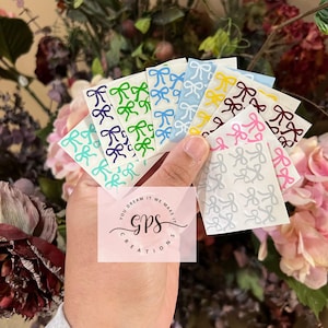 May include: Assortment of ten sheets of small, white, adhesive, vinyl stickers with a repeating pattern of gray bows. Each sheet has a different colored bow, including teal, green, blue, yellow, red, pink, and purple.