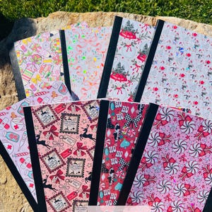 May include: A collection of notebooks with various festive designs. Patterns include Christmas ornaments, candy canes, reindeer, and gift boxes. The notebooks have black spines and are arranged on a stone surface with green grass in the background.