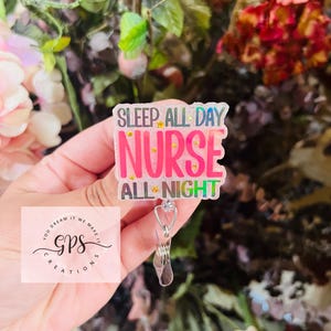 May include: A clear badge reel with the text "SLEEP ALL DAY NURSE ALL NIGHT" in pink, white, and green. The badge reel has a silver clip and a retractable cord. The background is blurred with colorful flowers and foliage.