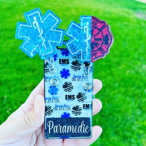 May include: A blue and red glitter badge holder with the words "EMS love" and "Paramedic" on it. The badge holder has a design of stars and the words "Saving lives one at a time".