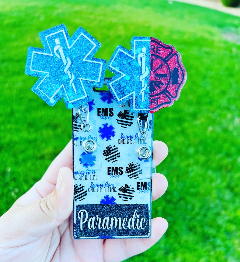 May include: A blue and red glitter badge holder with the words "EMS love" and "Paramedic" on it. The badge holder has a design of stars and the words "Saving lives one at a time".